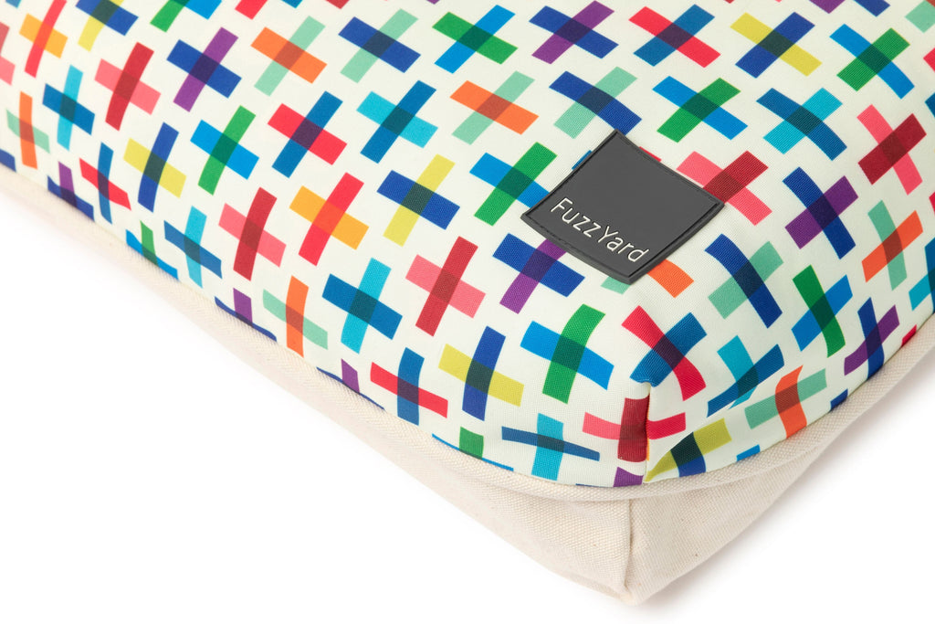 A close-up of a rectangular cushion with a colorful geometric pattern of intersecting crosses on a cream background, finished with a solid beige fabric binding. A black fabric brand tag reading 'Fuzzy Yard' is sewn onto the side.