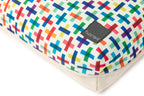 A close-up of a rectangular cushion with a colorful geometric pattern of intersecting crosses on a cream background, finished with a solid beige fabric binding. A black fabric brand tag reading 'Fuzzy Yard' is sewn onto the side.