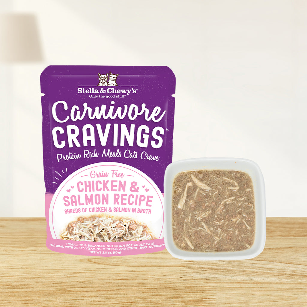 A pouch of Stella & Chewy's Carnivore Cravings grain-free chicken and salmon Recipe wet cat food next to a small white square bowl filled with the actual shredded chicken and salmon meat in broth