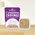 A pouch of Stella & Chewy's Carnivore Cravings grain-free chicken and tuna ecipe wet cat food next to a small white square bowl filled with the actual shredded tuna and visible chicken chunks in broth