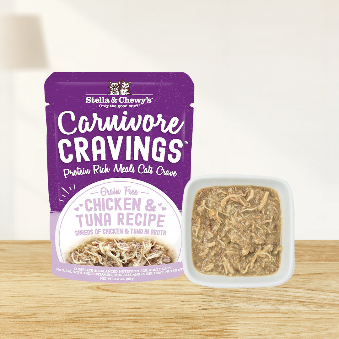 A pouch of Stella & Chewy's Carnivore Cravings grain-free chicken and tuna ecipe wet cat food next to a small white square bowl filled with the actual shredded tuna and visible chicken chunks in broth
